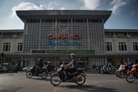 Many motorcyclists ride in front of the station building. One of them looks directly into the camera