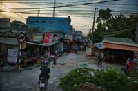 A street runs through small stores. A moped drives through the narrow street. The locals sit on the edge, working and cooking